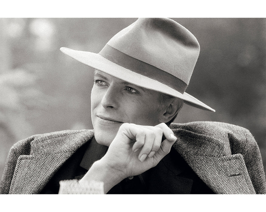 A black and white image of David Bowie in a tweed jacket and wide-brimmed hat, captured by Terry O'Neill 